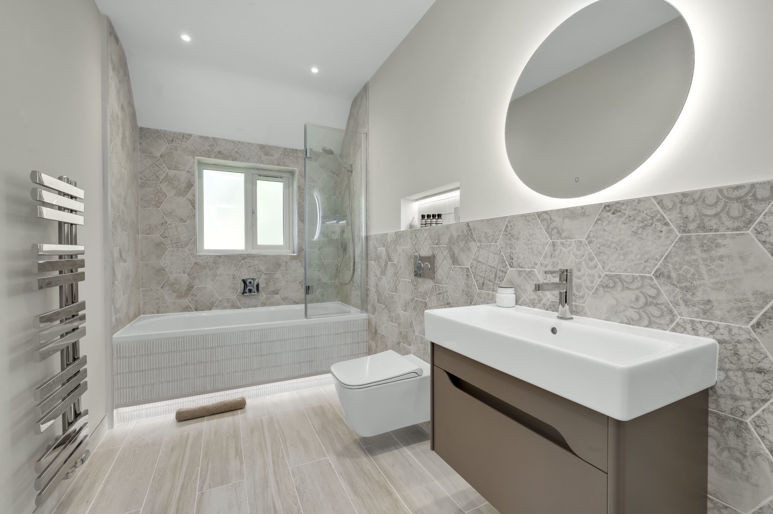 Modern bathroom with patterned wall tiles, a bath with glass screen, wall-hung toilet and illuminated mirror