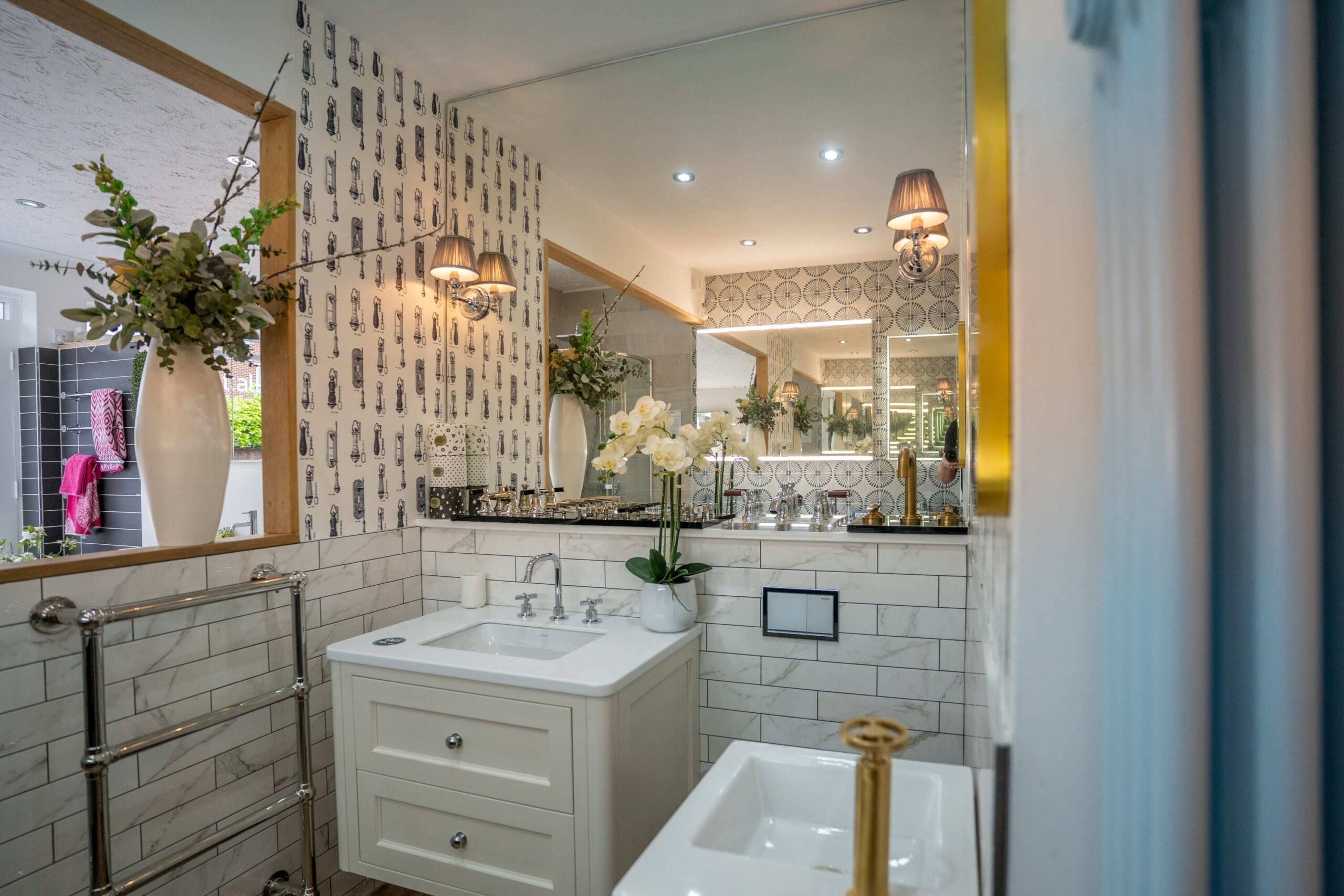 Traditional bathroom showroom display with a white vanity unit, patterned wallpaper and illuminated mirrors