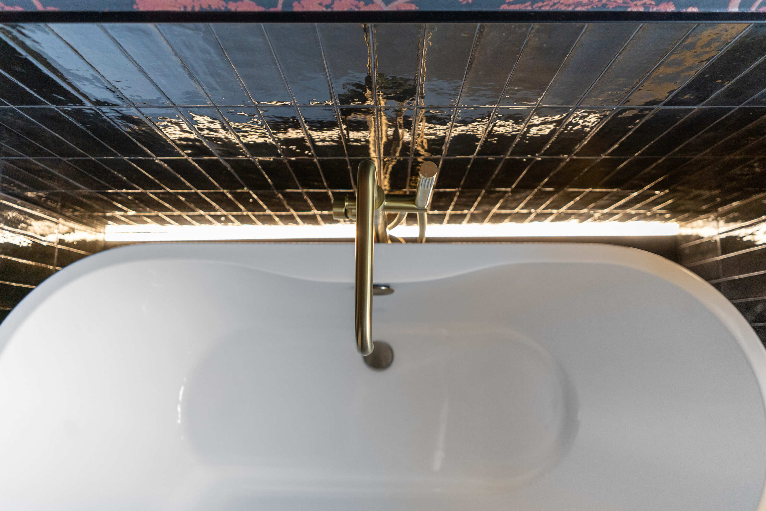 Overhead view of a white freestanding bath with a brushed brass tap and glossy black wall tiles